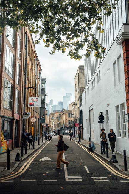 A street view in Shoreditch featuring a row of multi-storey buildings with a mix of brick and light-colored facades, large windows, and decorative architectural details. In the foreground, a white mov